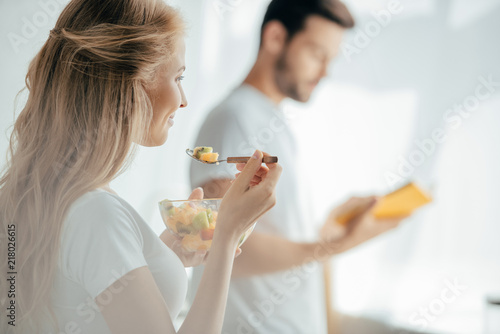selective focus of smiling pregnant woman eating fruits salad while husband standing at counter in kitchen
