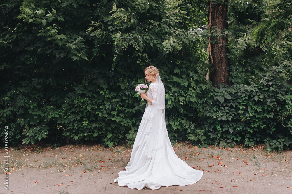 A beautiful bride in a lace dress is standing with a bouquet in her hands against the backdrop of summer nature and green foliage. Wedding portrait of a curly bride. A beautiful bride in a lace dress is standing with a bouquet in her hands against the backdrop of summer nature and green foliage. Wedding portrait of a curly bride.
