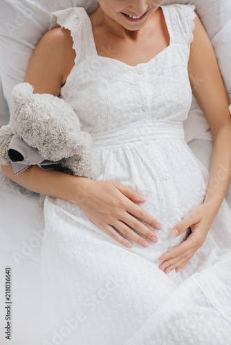 overhead view of smiling pregnant woman in white nightie with teddy bear resting on sofa at home