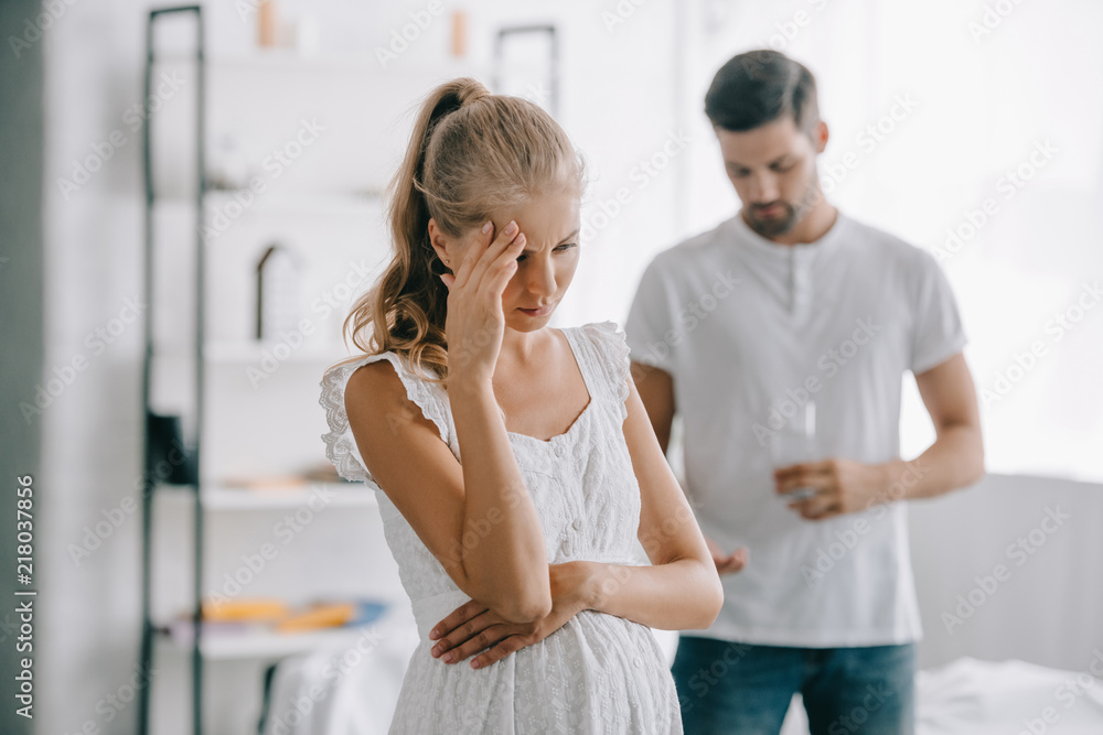 Obraz premium pregnant woman in white nightie having headache while husband with medicines and glass of water standing behind at home