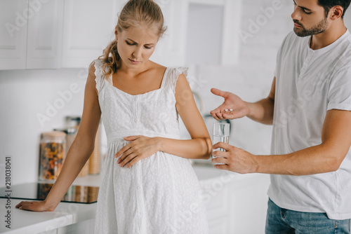 pregnant woman in white nightie having discomfort while husband giving medicines and glass of water to her at home