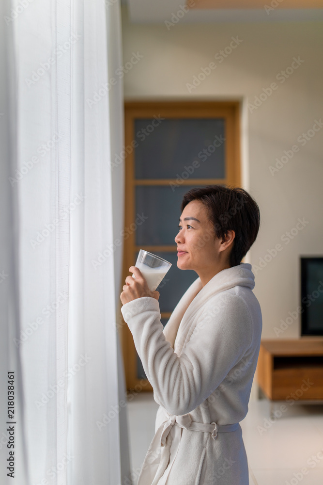 Asian woman drinking a glass of milk in living room and looking outside window with smile face