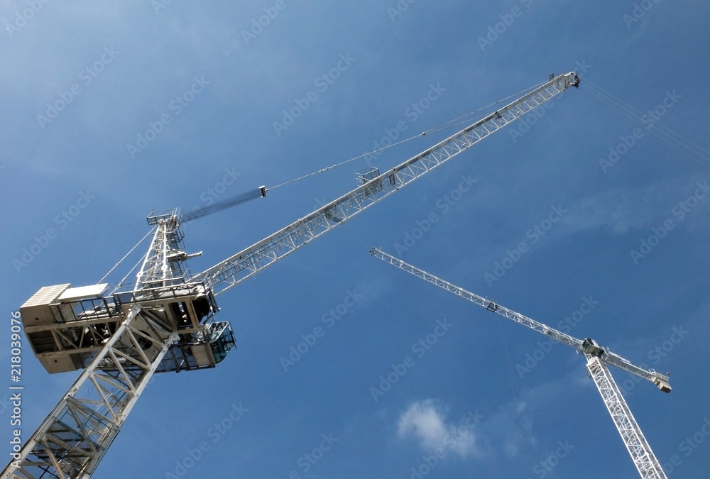 a perspective view of two tall white construction cranes on a building site against a blue sky with clouds