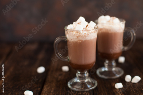 Two glasses with Hot chocolate garnished with whipped cream, marsmallow and cocoa powder.