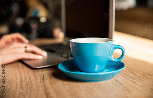 Crop of woman sitting in cafe and working with laptop  press buttons. Blue glass of coffee near on wooden table. Freelancer working at loft space  having lunch or breakfast alone.