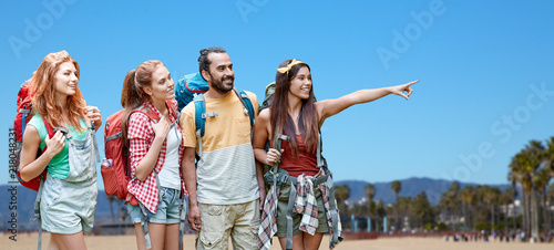 travel, tourism, hike and adventure concept - group of smiling friends with backpacks pointing finger to something over venice beach background in california