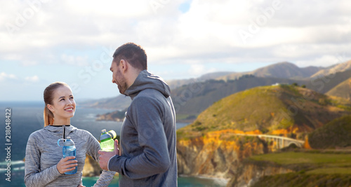 fitness, sport and people concept - smiling couple with bottles of water over bixby creek bridge on big sur coast of california background