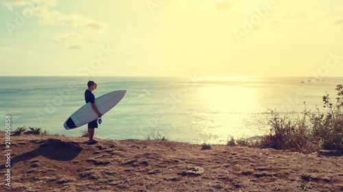 Surfer atay at top of the cliff near sand beach, looking the waves at sunset time.Dreamland beach area,Bali island,Indonesia