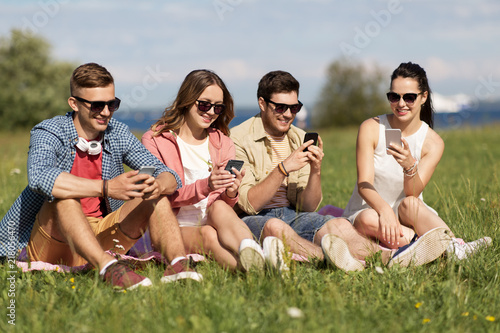 friendship, technology and leisure concept - group of smiling friends with smartphones sitting on grass in summer