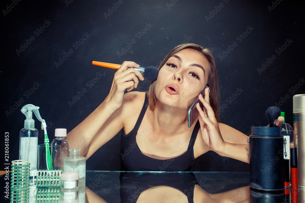 Obraz premium Beauty woman applying makeup. Beautiful girl looking in the mirror and applying cosmetic with a big brush. Morning, make up and human emotions concept. Caucasian model at studio
