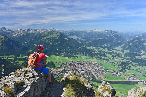 Female hiker at the summit of Rubihorn mountain enjoying the view to the village Oberstdorf and the Allgau Alps. Bavaria, Germany. Alpine landscape with rocky mountains, forests and blue sky.