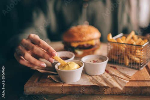 French fries and ketchup in basket with flare light. Fast food, people and eating concept. Close up of hand with dipping french fries into ketchup bowl on wooden table. Selective focus.