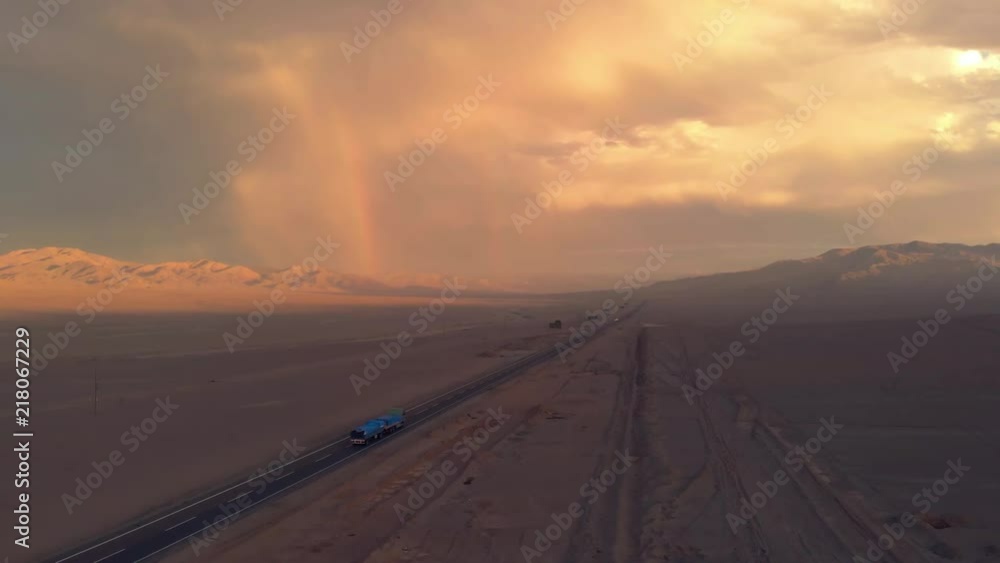 Aerial desert sunset with rainbow, road and colorful sky. Atacama Chile. Drone rising up filming road with truck driving forward during beautiful evening. Empty vast landscape with warm color grade. Aerial desert sunset with rainbow, road and colorful sky. Atacama Chile. Drone rising up filming road with truck driving forward during beautiful evening. Empty vast landscape with warm color grade.