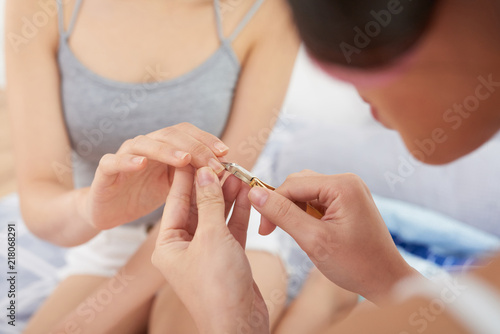 Unrecognizable female using cuticle nipper to help friend with manicure while sitting in bedroom together photo