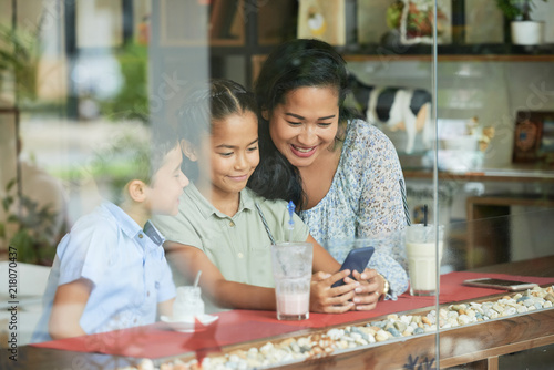 Asian woman and children smiling and browsing smartphone while sitting behind window glass in cafe