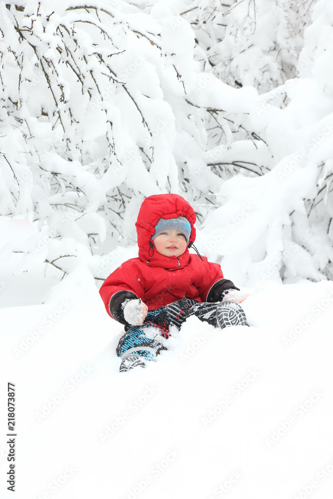 Cute toddler boy in winter suit sitting on a big snowdrift, surrounded by snow-covered trees. Snowy winter. Copy space
