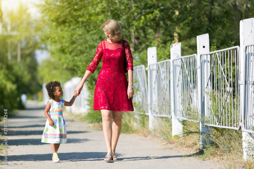 Naklejka premium A white woman is walking along the street with a black girl. A European mother with an African daughter in summer dresses is walking with her hand.