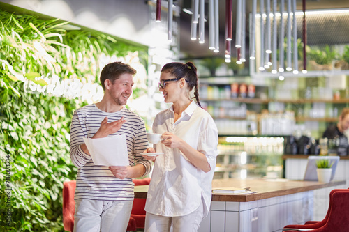 Stylish woman with coffee cup and man with papers standing in cafeteria and chatting in leisure