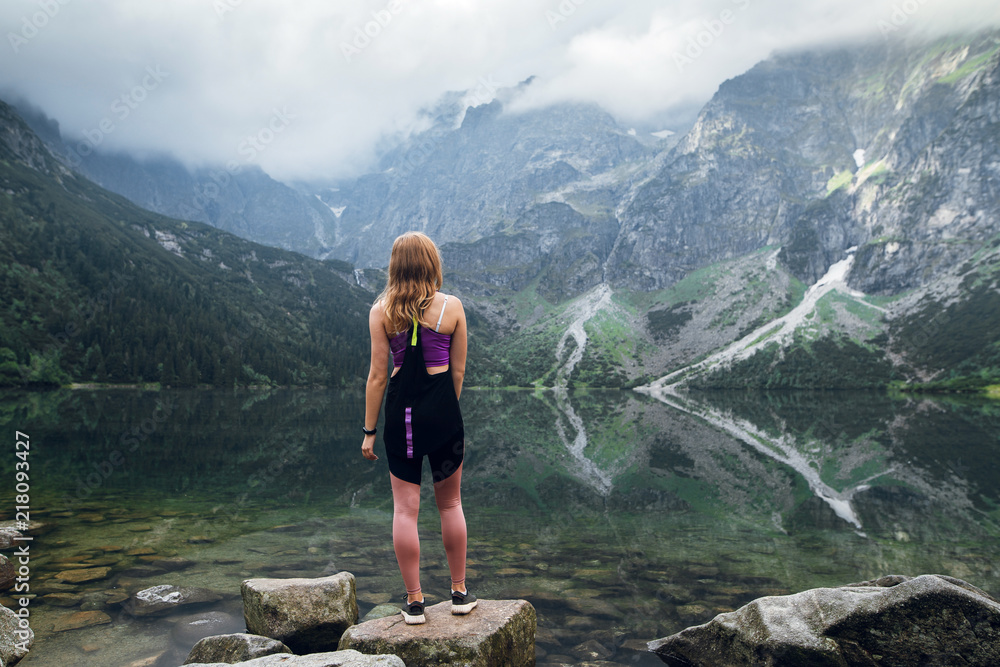 Naklejka premium Rear view shot of young fit woman in sportswear looking on the scenic green hills and mountains in dark clouds on the Marine Eye lake, High Tatras, Zakopane, Poland. Foggy day