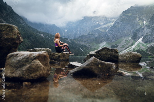 Young blonde fit and sporty woman in sportswear sitting on the rock on the shore and admiring beautiful view of green hills and mountains on Morskie Oko lake, High Tatras, Zakopane, Poland. © WellStock