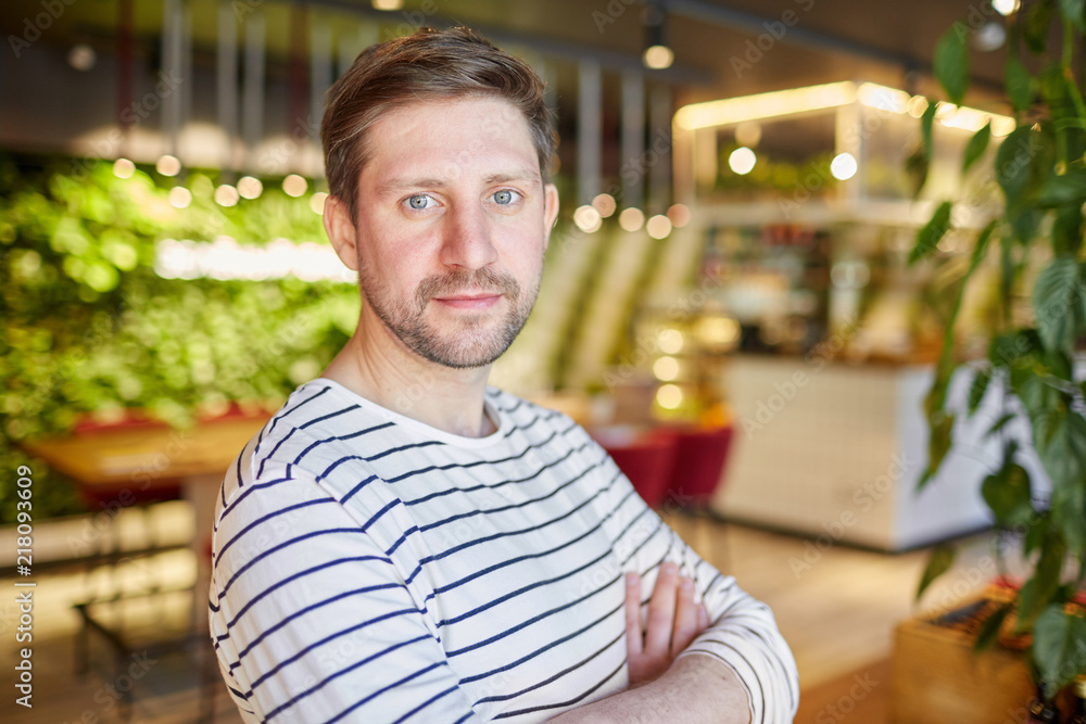 Obraz premium Handsome unshaven casual man in striped shirt standing with arms crossed and looking at camera in light modern interior