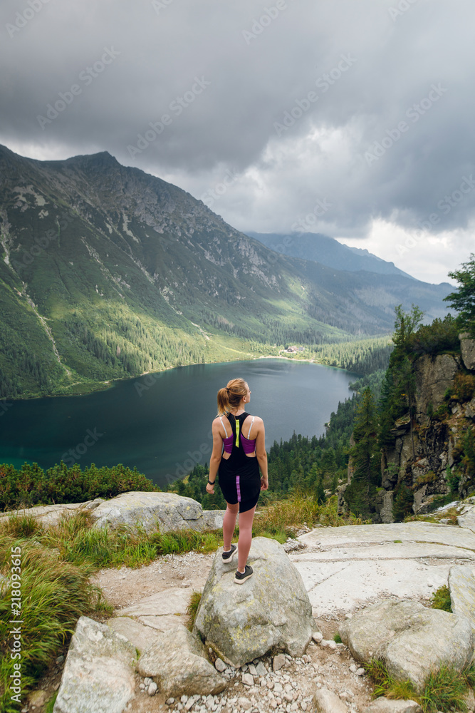 Back view of young blonde woman in sportswear standing on the rock and enjoying beautiful view of green mountains on Morskie Oko lake, High Tatras, Zakopane, Poland. Top view