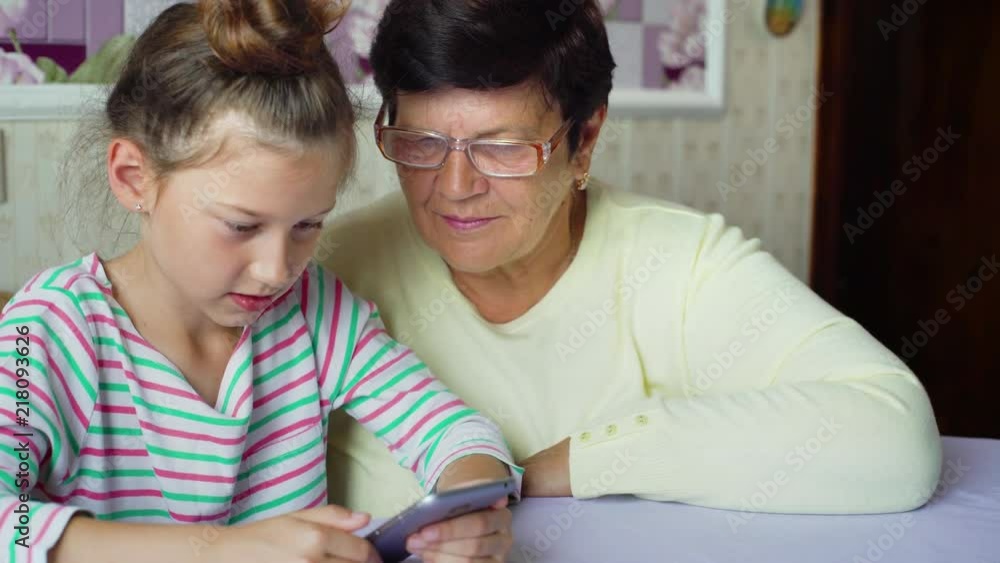 Young cute granddaughter teaching grandmother how to use smartphone at home. Small girl and elderly woman have leisure time together. Family, relation, communication, technology, education concept Young cute granddaughter teaching grandmother how to use smartphone at home. Small girl and elderly woman have leisure time together. Family, relation, communication, technology, education concept