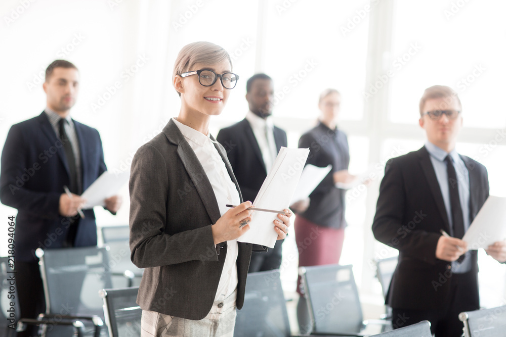 Group of coworkers holding papers with notes and standing in hall with chair having meeting Group of coworkers holding papers with notes and standing in hall with chair having meeting