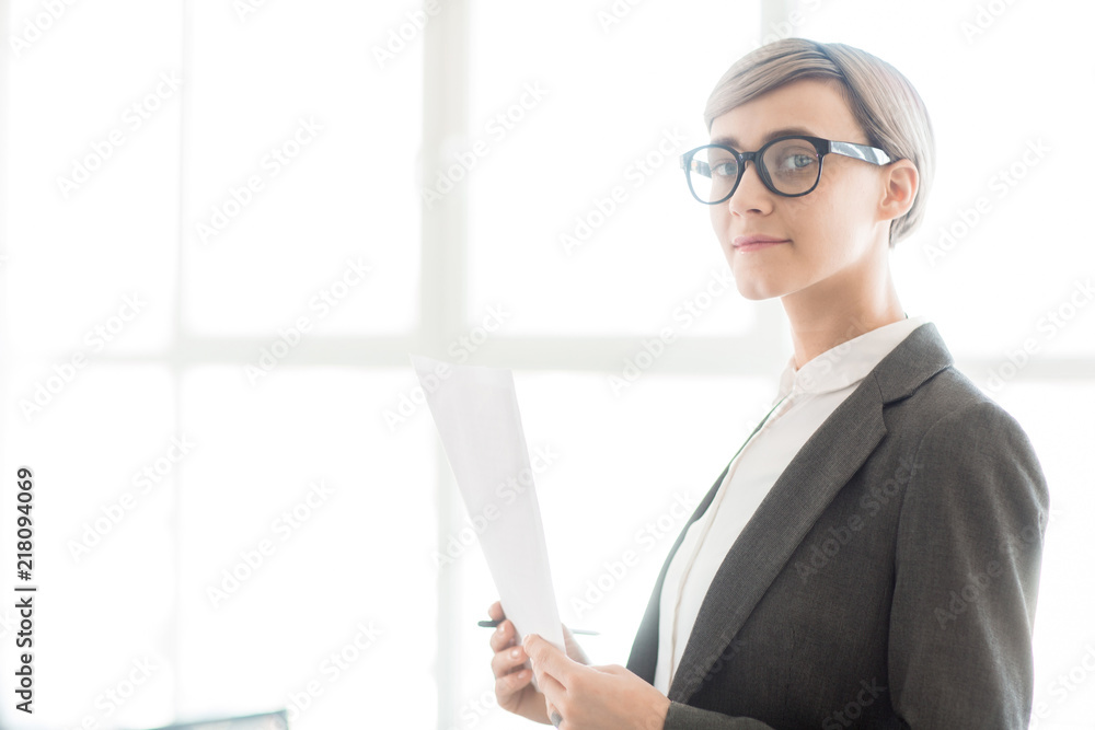 Adult professional woman with trendy short hairstyle wearing jacket and glasses holding paper and looking at camera Adult professional woman with trendy short hairstyle wearing jacket and glasses holding paper and looking at camera