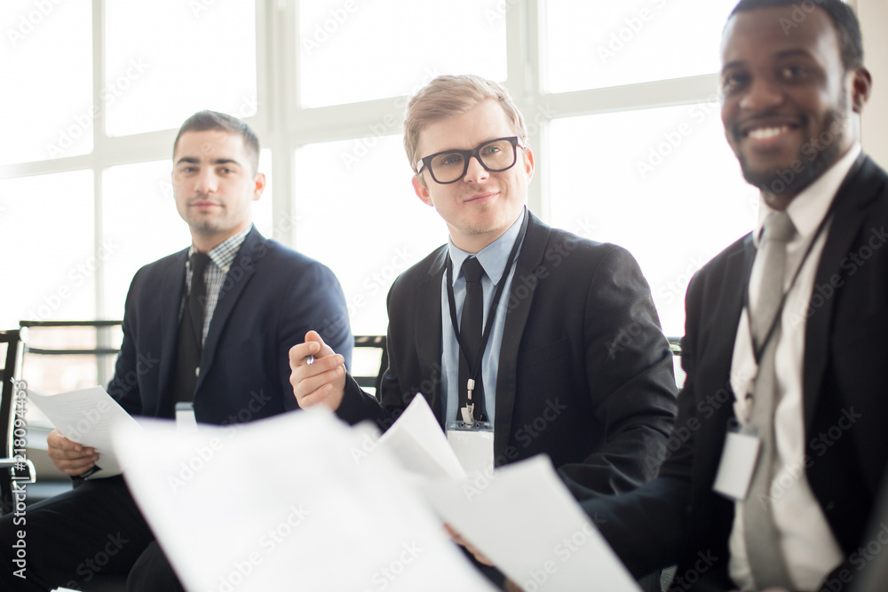 Fototapeta premium Adult multiracial men in suits sitting with papers in hands and communicating on business meeting