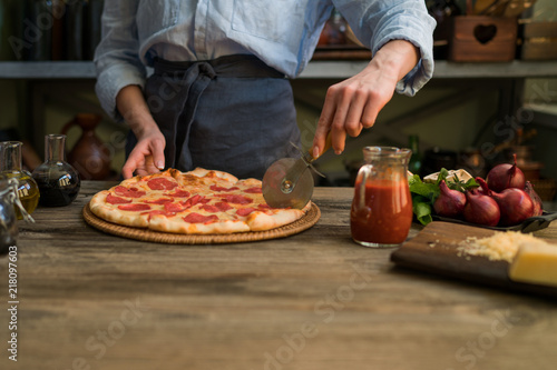 Woman cutting fresh baked homemade pizza on rustic kitchen background. Cut into slices delicious pizza with mushrooms and ham. Cheese and tomatoes on wooden table. Healthy foods  cooking concept.