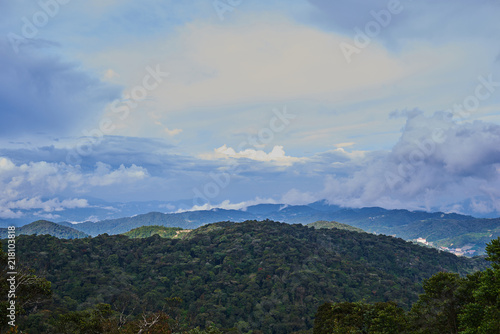 Mountain hill covered with forest on foreground and curly clouds on background with foggy valley with village in between. Mountains during sunrise. Beautiful natural landscape in the summer time..