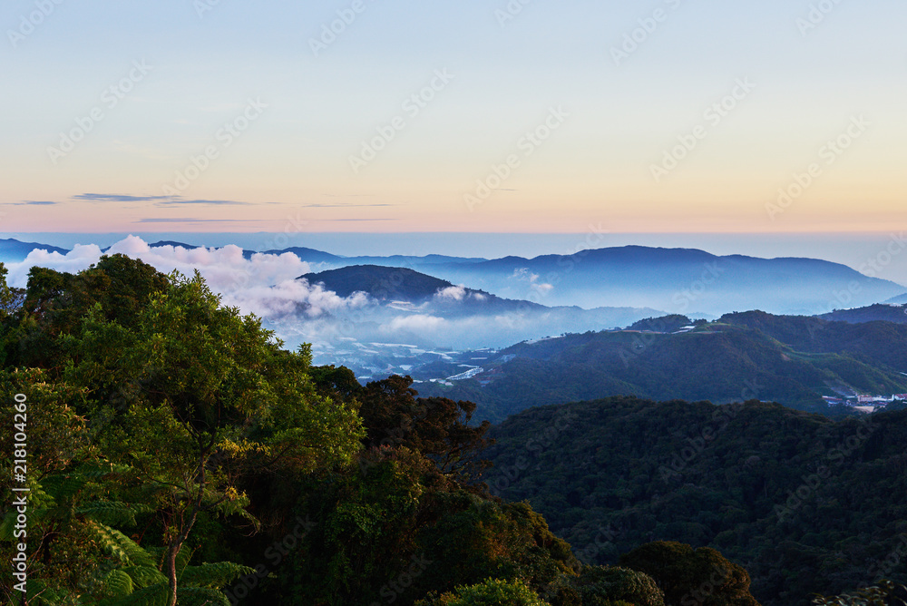 Mountain hill covered with forest on foreground and curly clouds on background with foggy valley with village in between. Mountains during sunrise. Beautiful natural landscape in the summer time..