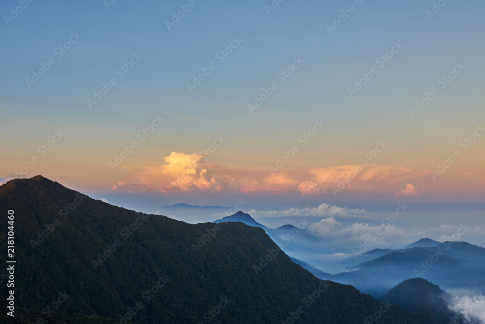 Hills in valley between the mountains with fog and curly clouds illuminated by the rising sun.