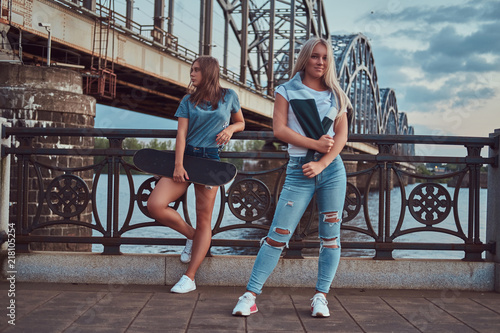 Two beautiful hipster girls standing with skateboard against a bridge.