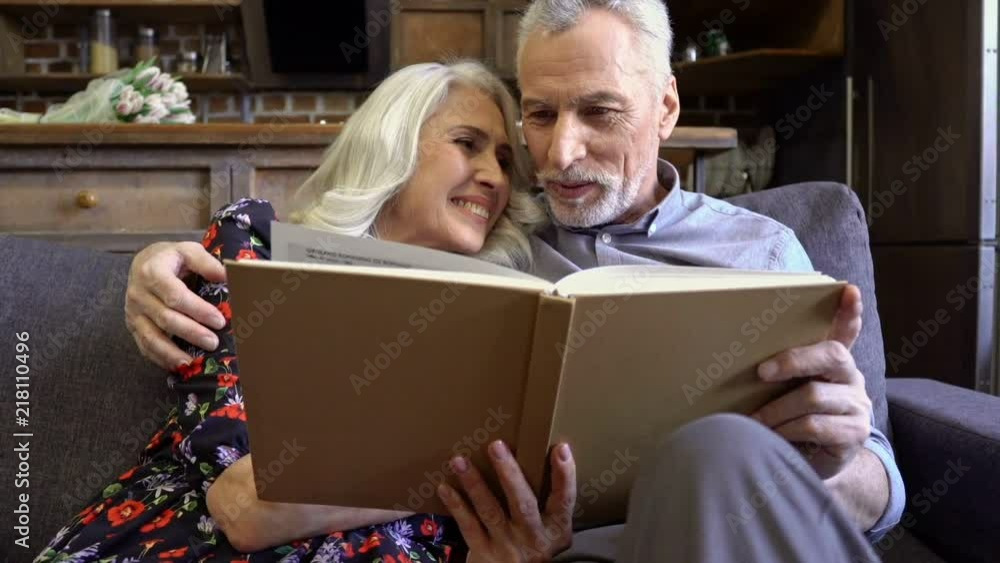 Smiling lovely elderly couple reading book and talking together while sitting together on sofa at home Smiling lovely elderly couple reading book and talking together while sitting together on sofa at home