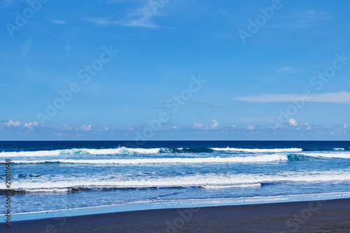 Black sand volcanic beach with waves and deep blue ocean. Summer tourism and holiday concept. Blue sea water and blue sky. View amazing nature background. Exsotic landscape with copy space..