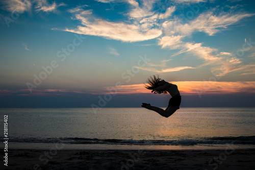 junge Ballerina vollzieht einen Sprung am Strand mit Meer und einem perfekten Sonnenuntergang im Hintergrund
