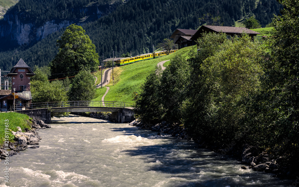 Picturesque alpine scene with a river, some typical wooden chalets and a yellow train coming down the mountain. Grindelwald, Bernese Oberland, Switzerland