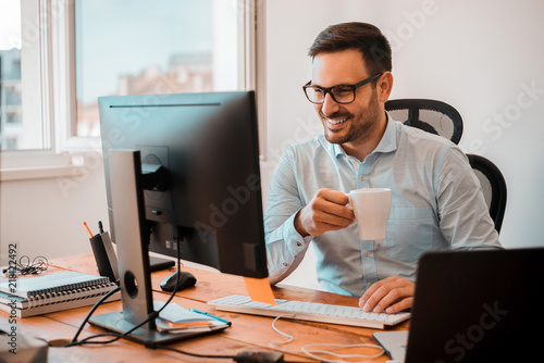 Handsome man drinking coffee while looking at computer monitor at home office. photo