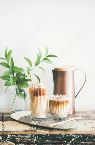 Iced coffee in glasses with milk and straws on board over rustic wooden table, white wall, jug and plant branch in vase at background, copy space. Summer refreshing beverage, ice coffee drink concept