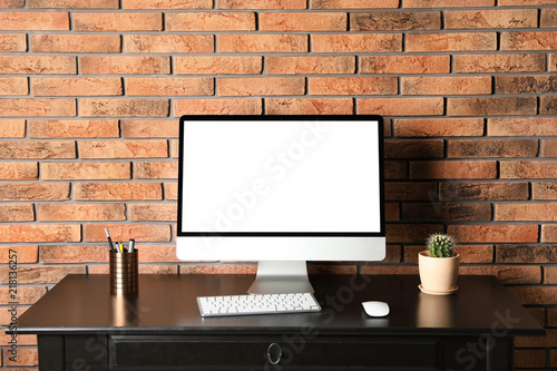 Modern computer monitor on desk against brick wall, mock up with space for text