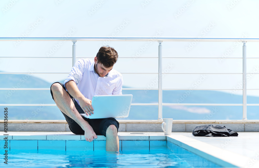 Handsome young man relaxing and working on laptop computer at home balcony while looking sea