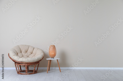 Comfortable papasan chair and lamp on table near color wall with space for text. Interior element photo
