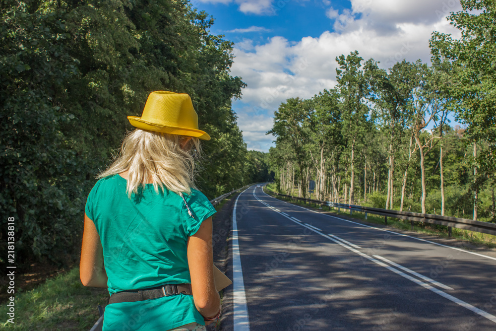 hitchhiking journey travel girl concept back to camera with bright clothes green t-short and yellow hat near empty concrete car road between green trees in nature scenery colorful summer environment