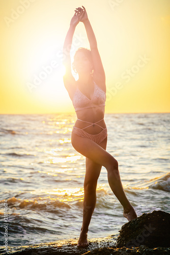 silhouette of a girl in a swimsuit stands on a stone beach, sunbathing at dawn, raising her hands.
