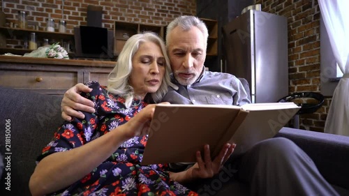 Cheerful elderly elegant couple sitting together on sofa and reading book at home while man kissing woman photo