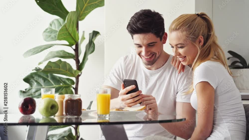 Handsome young lovers having breakfast while sitting at the table in a kitchen at home and using smartphone Handsome young lovers having breakfast while sitting at the table in a kitchen at home and using smartphone
