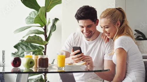 Handsome young lovers having breakfast while sitting at the table in a kitchen at home and using smartphone photo