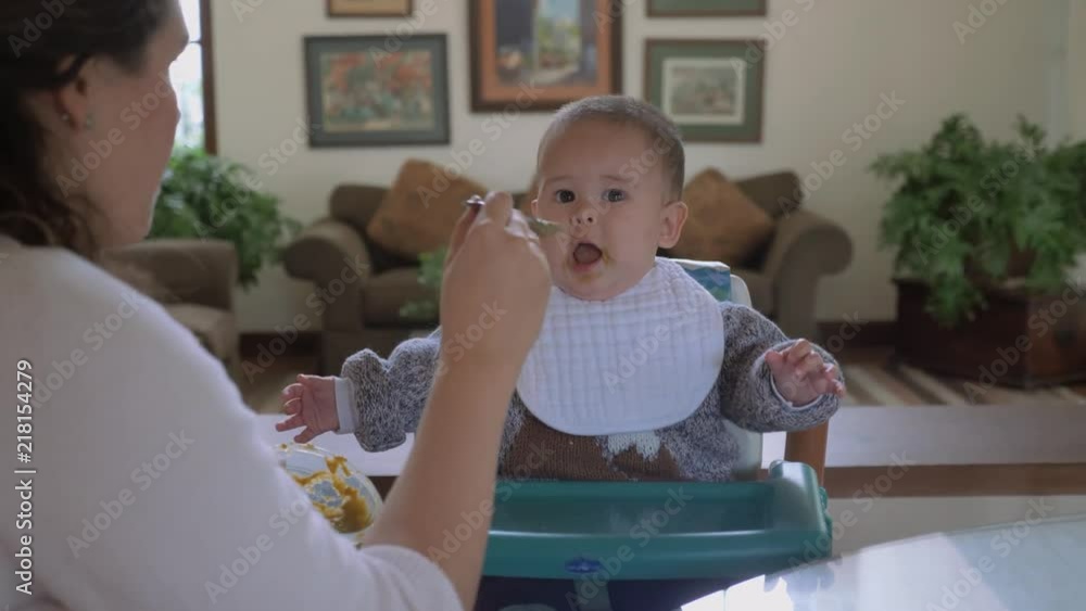Mother feeding his baby boy with green vegetables pap in living room at home. Winter time clothing. Child routine, day-to-day activities. Pan shot. Mother feeding his baby boy with green vegetables pap in living room at home. Winter time clothing. Child routine, day-to-day activities. Pan shot.